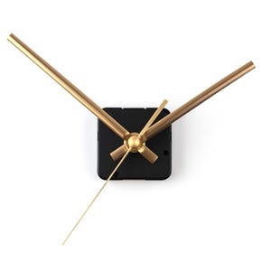 May include: A modern clock mechanism with a black square base and gold-colored hands. The hour and minute hands are long and slender, with a delicate second hand. The clock hands are positioned to indicate the time.
