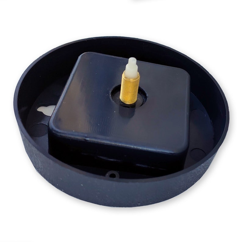May include: A black clock movement with a gold-coloured stem and a white plastic knob. The movement is in a black plastic housing.
