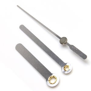 May include: Three silver clock hands with gold accents. The hands are of different lengths and are designed for a clock face.