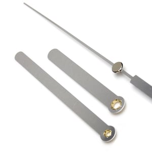May include: Three silver clock hands, one long, one medium, and one short. The hands have gold-coloured metal inserts at the ends.