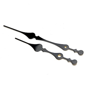 May include: A pair of black clock hands with a decorative design. The hands are made of metal and have a gold-colored accent.
