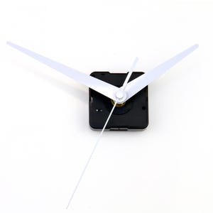 May include: Clock mechanism with a black square body and white hands. The hour and minute hands are wide and flat, and the second hand is a thin, straight line. The clock mechanism is set against a white background.