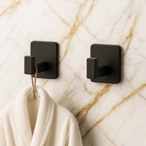 Puede incluir: Dos perchas de pared negras mate con placa trasera cuadrada y gancho rectangular. Una bata blanca cuelga de una percha. Las perchas están montadas en una pared con diseño de mármol, ofreciendo un diseño moderno y minimalista para el baño o dormitorio.