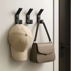 Könnte beinhalten: Drei schwarze Metallhaken an einer weißen Wand. Eine beige Baseballkappe und eine taupefarbene Handtasche hängen an den Haken. Die Haken haben ein modernes, minimalistisches Design, ideal für Flur oder Schlafzimmer.