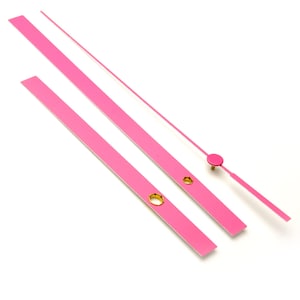 Peut inclure: Trois aiguilles d'horloge roses, une longue, une moyenne et une courte, avec des attaches en métal doré. Les aiguilles sont isolées sur un fond blanc.