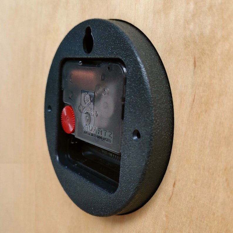 May include: Close-up of the back of a black clock with a quartz movement. The clock has a red knob and a clear plastic cover over the movement. The text "QUARTZ" is visible on the movement.