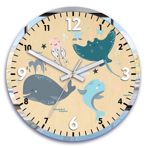 May include: A round wooden clock with a light brown background and blue accents. The clock face features a whale, a jellyfish, a stingray, and a narwhal, all with smiling faces. The numbers 1 through 12 are printed in black around the clock face.