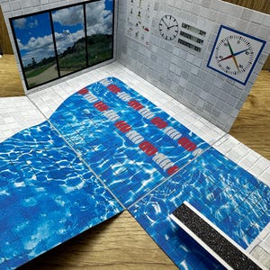 Könnte beinhalten: Miniatur-Papier-Schwimmbad-Diorama mit blauem Wassermuster, rot-weißen Bahnenmarkierungen und einem Fenster mit Landschaft. Die Wände sind gefliest, mit einer Uhr und einem Temperaturmessgerät. Das Diorama ist offen und auf einer Holzoberfläche ausgestellt.