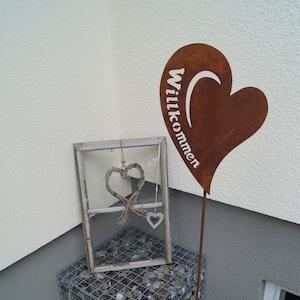 May include: A rusty metal heart-shaped sign with the word "Willkommen" written on it. The sign is attached to a metal pole and is standing in front of a white wall. There is a small window frame with a woven heart hanging inside.