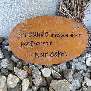 Puede incluir: Un letrero de metal oxidado con forma ovalada blanca y el texto "Freunde m&uuml;ssen nicht perfekt sein. Nur echt." colgado de una cuerda.