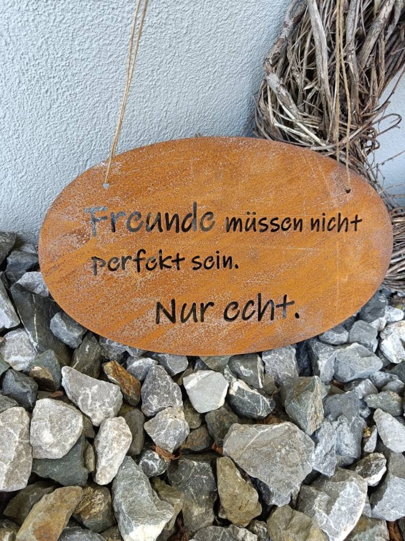 Puede incluir: Un letrero de metal oxidado con forma ovalada blanca y el texto "Freunde m&uuml;ssen nicht perfekt sein. Nur echt." colgado de una cuerda.