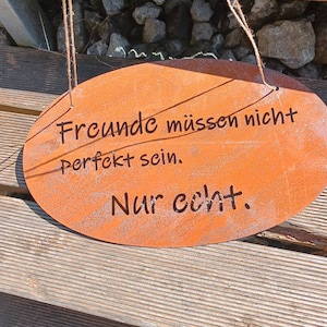 Puede incluir: Un letrero ovalado de metal oxidado de color naranja con el texto "Freunde m&uuml;ssen nicht perfekt sein. Nur echt." El letrero est&aacute; colgado de una superficie de madera con cuerda.