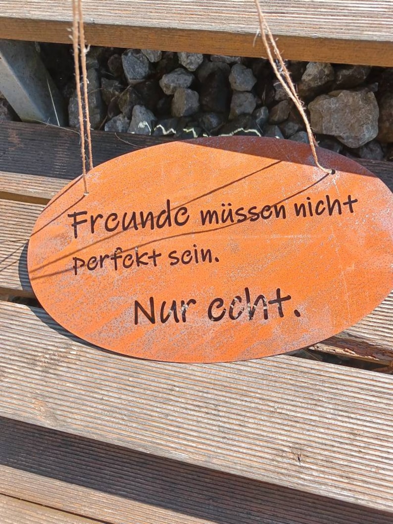Puede incluir: Un letrero ovalado de metal oxidado de color naranja con el texto "Freunde m&uuml;ssen nicht perfekt sein. Nur echt." El letrero est&aacute; colgado de una superficie de madera con cuerda.