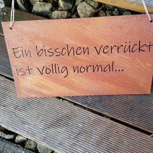Può includere: Un cartello di metallo arrugginito con il testo "Ein bisschen verrückt ist völlig normal..." appeso su una superficie di legno.
