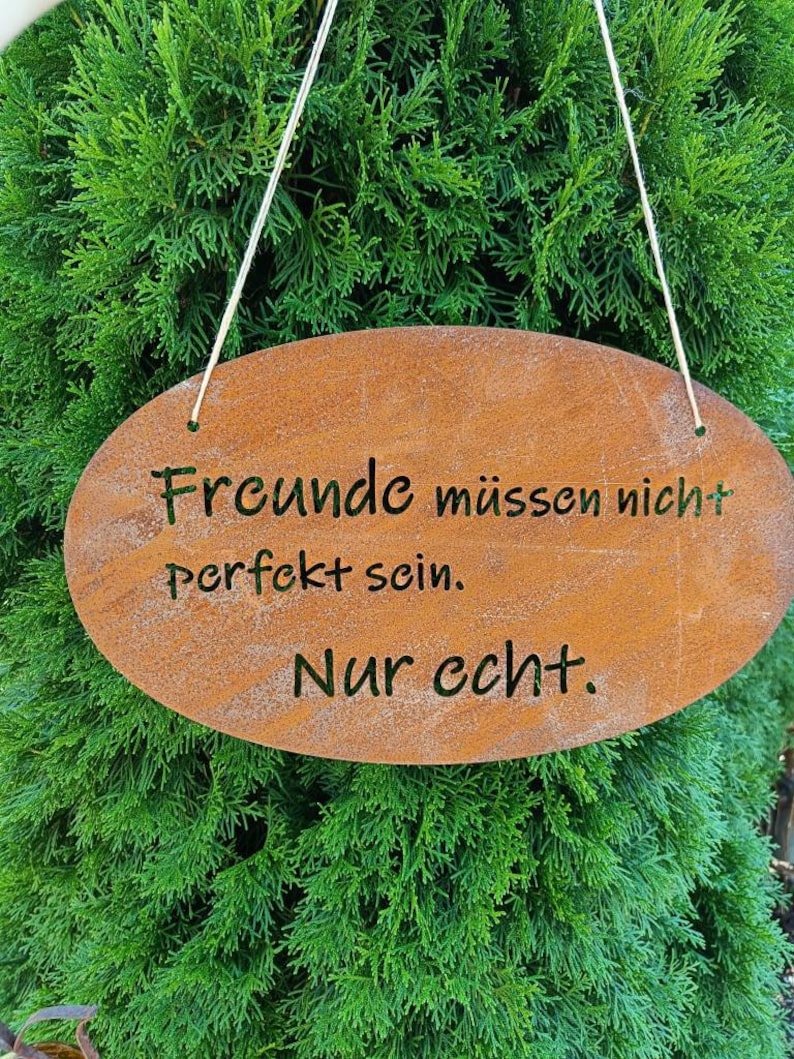 Puede incluir: Un letrero ovalado de metal marr&oacute;n oxidado con el texto "Freunde m&uuml;ssen nicht perfekt sein. Nur echt." colgado de una cuerda blanca sobre un fondo de arbusto verde.