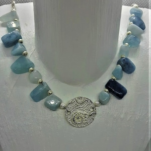 Könnte beinhalten: Eine silberne Halskette mit einem runden, filigranen Anhänger und blauen und grünen Edelsteinperlen.