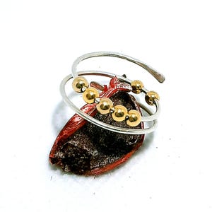 Könnte beinhalten: Ein silberfarbener Wickelring mit goldfarbenen Perlen. Der Ring hat ein Spiraldesign mit mehreren goldenen Perlen. Der Ring wird auf einer dunkelroten und braunen strukturierten Oberfläche vor weißem Hintergrund präsentiert.