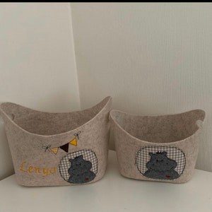 Op de afbeelding: Twee beige wollen opbergmanden met een nijlpaard ontwerp. De grotere mand heeft de naam "Lenya" geborduurd. Beide manden hebben een grijs en wit geruit patroon en een gele, bruine en zwarte vlaggenlijn.