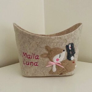 Op de afbeelding: Een beige wollen opbergmand met een hert en konijn applique. De mand heeft de naam "Maila Luna" geborduurd op de voorkant in roze draad.