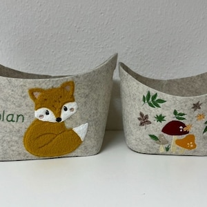 Op de afbeelding: Twee lichtgrijze vilten opbergmanden. Eén heeft een vossenapplicatie en de naam "Nolan" in groen geborduurd. De andere heeft geborduurde paddenstoelen en bladeren. Deze decoratieve manden zijn ideaal voor het organiseren van spullen.