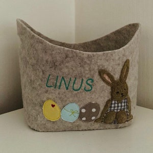 Op de afbeelding: Een grijze vilt paasmand met een konijnenapplicatie, drie vilt paaseieren en de naam "LINUS" geborduurd op de voorkant.