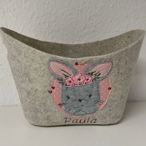 Op de afbeelding: Een grijze wollen opbergmand met een roze en grijze konijn geborduurd op de voorkant. Het konijn draagt een roze bloemenkrans en de mand is gepersonaliseerd met de naam "Paula".