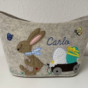 Op de afbeelding: Een grijze vilt paasmand met een konijn dat een karretje trekt met twee paaseieren. Het konijn is bruin met een blauw-wit geblokte sjaal. De eieren zijn versierd met witte stippen en een geel lint. De mand is gepersonaliseerd met de naam "Carlo".