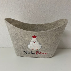Op de afbeelding: Een lichtgrijze vilten mand met een wit geborduurd kippenontwerp en de woorden "Frohe Ostern" in zwart en rood. De mand heeft een gebogen, open bovenkant en is geschikt voor paasdecoraties.