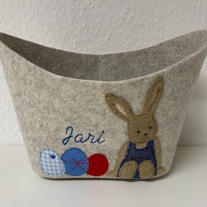 Op de afbeelding: Een grijze vilt paasmand met een konijn en drie paaseieren. Het konijn draagt een blauwe denim outfit. De mand is gepersonaliseerd met de naam "Jari".