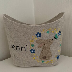 Op de afbeelding: Een beige wollen opbergmand met een geborduurd konijn- en vlinderontwerp. De mand heeft een gele en groene bloemenkrans met blauwe bloemen. De naam Henri is geborduurd op de voorkant van de mand.