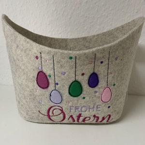 Op de afbeelding: Een grijze wollen paasmand met hangende roze, paarse, groene en lichtpaarse eieren en de tekst "Frohe Ostern" geborduurd op de voorkant.