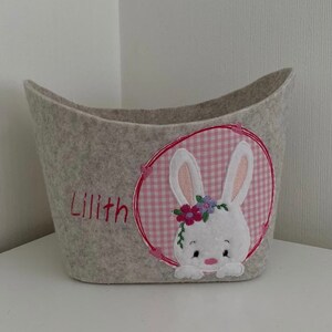 Op de afbeelding: Een grijze vilt opbergmand met een roze en witte geruite konijnenapplicatie. De mand is geborduurd met de naam "Lilith".