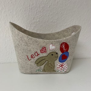 Op de afbeelding: Een grijze wollen opbergmand met een konijn- en ballonontwerp. De mand heeft de naam "Lea" geborduurd op de voorkant.