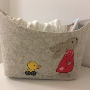 Op de afbeelding: Een grijze wollen opbergmand met een konijn- en eend-borduursel. De mand is gevuld met witte katoenen luiers.
