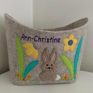 Op de afbeelding: Een grijze wollen opbergmand met een konijn, bloemen en groene bladeren geborduurd op de voorkant. De mand is gepersonaliseerd met de naam "Ann-Christine".