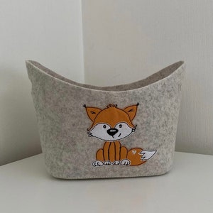 Op de afbeelding: Een grijze wollen opbergmand met een vos-ontwerp. De vos is oranje met zwarte ogen en een witte neus. De mand is bootvormig.
