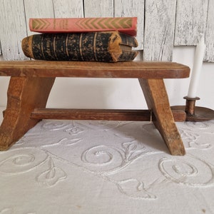 Pode incluir: Um pequeno banquinho de madeira com um design simples, com um topo plano e duas pernas inclinadas. O banquinho é pintado em uma cor de madeira natural e tem uma aparência rústica. Dois livros e um castiçal estão colocados em cima do banquinho.