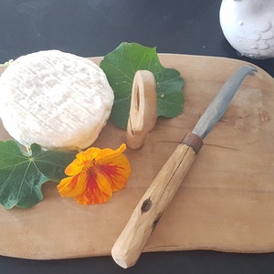 Peut inclure: Un fromage rond et blanc sur une planche à découper en bois, accompagné d'un couteau à fromage, d'un marqueur à fromage en bois, de feuilles vertes et d'une fleur orange. Le couteau à fromage a un manche en bois et une lame incurvée.