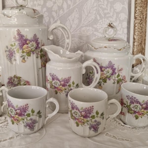 Peut inclure: Un service à thé en porcelaine vintage avec un motif floral. L'ensemble comprend une théière, un sucrier, une crémière et six tasses. La porcelaine blanche est ornée de fleurs violettes, jaunes et vertes. L'ensemble est présenté sur une nappe en dentelle blanche.
