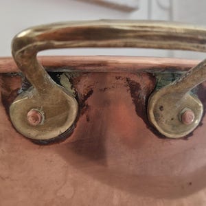 Peut inclure: Gros plan sur une casserole en cuivre avec une poignée en laiton. La casserole a une forme arrondie avec une finition en cuivre brillant. La poignée en laiton est incurvée et fixée à la casserole avec des rivets en laiton. La casserole semble être vintage.