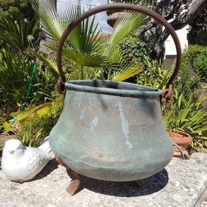 Pode incluir: Uma panela de cobre antiga grande com uma alça e três pernas. A panela é verde com a idade e tem um aspecto rústico e desgastado. Está sentada sobre uma superfície de pedra num jardim.