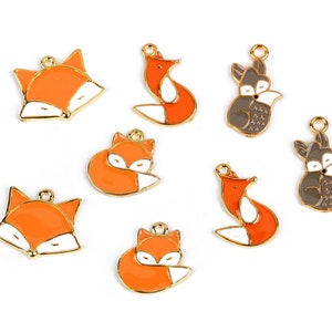 Könnte beinhalten: Eine Sammlung von Fuchs-Anhängern aus Emaille. Die Anhänger zeigen orange, weiße und graue Füchse mit goldfarbenen Umrissen. Einige Füchse liegen, andere stehen oder haben den Kopf gedreht.