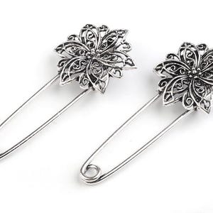 Könnte beinhalten: Zwei silberne Sicherheitsnadeln mit dekorativen Blumendesigns. Die Blumen sind filigran und haben ein filigranes Muster.