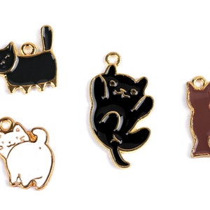 Könnte beinhalten: Vier goldfarbene Metall-Katzenanhänger mit Emaille-Lack. Ein Anhänger ist schwarz-weiß, einer ist schwarz, einer ist braun und einer ist weiß.