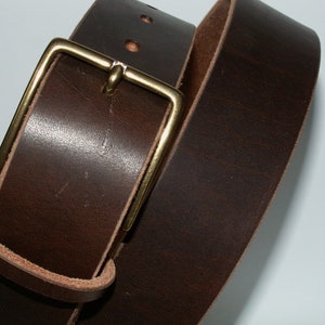May include: Close-up of a dark brown leather belt with a brass buckle. The belt has a rectangular buckle and several holes for adjustment. The leather appears to be thick and durable, suggesting a high-quality accessory.