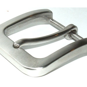 Könnte beinhalten: Eine silberne Metallschnalle mit glatter, gebürsteter Oberfläche. Die Schnalle hat eine rechteckige Form mit abgerundeten Ecken und einer gebogenen, federbelasteten Stange.