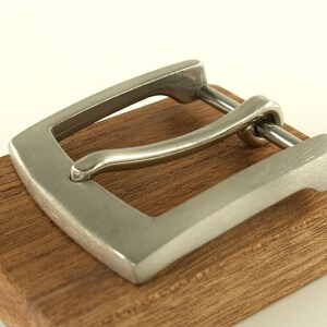 May include: A silver metal belt buckle with a smooth, brushed finish. The buckle is rectangular with rounded corners and has a simple, minimalist design.