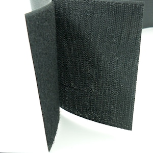 Könnte beinhalten: Nahaufnahme von schwarzem Klettverschlussmaterial. Das Material ist ein dunkler, strukturierter Stoff mit einer geschlungenen Oberfläche. Das Bild zeigt das gefaltete Material, das die Haken- und die Schlaufenseite zeigt.