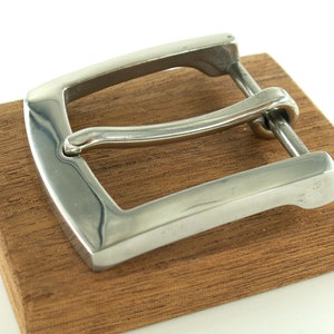 May include: A silver metal belt buckle with a smooth, polished finish. The buckle is rectangular with rounded corners and has a curved, spring-loaded tongue.