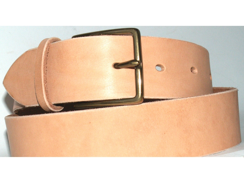 May include: A light tan leather belt with a brass buckle. The belt is rolled up, showing the buckle and the holes for the belt pin. The leather has a natural, unfinished look.